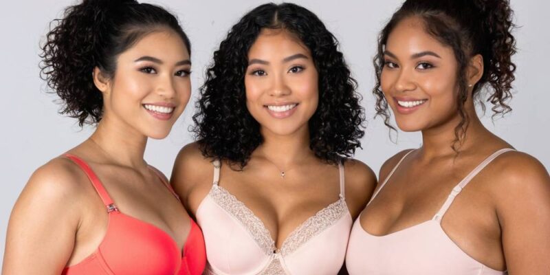 Three confident young women of diverse ethnicities standing together in a professional studio, each wearing properly fitted bras in coordinating coral, nude, and blush tones that celebrate their unique body types and support requirements. The woman on the left wears a coral push-up style bra with bright pink bottoms, the center woman wears a nude balconette bra with lace trim and matching briefs, and the woman on the right wears a soft blush wireless bralette with matching high-waisted shorts. All three display radiant smiles and relaxed confident postures with natural skin texture visible, demonstrating how proper bra fitting enhances comfort and self-assurance across different breast sizes, body shapes, and personal style preferences.